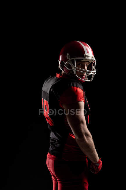 Young American football player in helmet standing against black background — Stock Photo