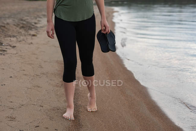 Baixa seção de mulher segurando chinelos na praia — Fotografia de Stock