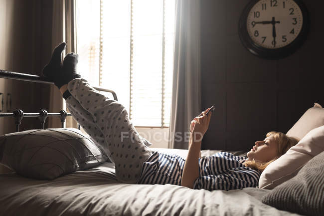 Вид сбоку женщины, пользующейся мобильным телефоном на кровати в спальне — стоковое фото