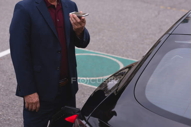 Sección media del hombre de negocios hablando en el teléfono móvil mientras carga el coche eléctrico - foto de stock