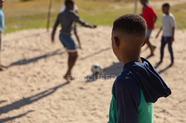 Menino olhando para as crianças jogando futebol no chão em um dia ensolarado — Fotografia de Stock
