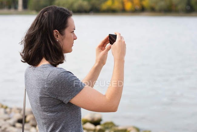 Hermosa mujer haciendo clic fotos con la cámara cerca de la orilla del río - foto de stock