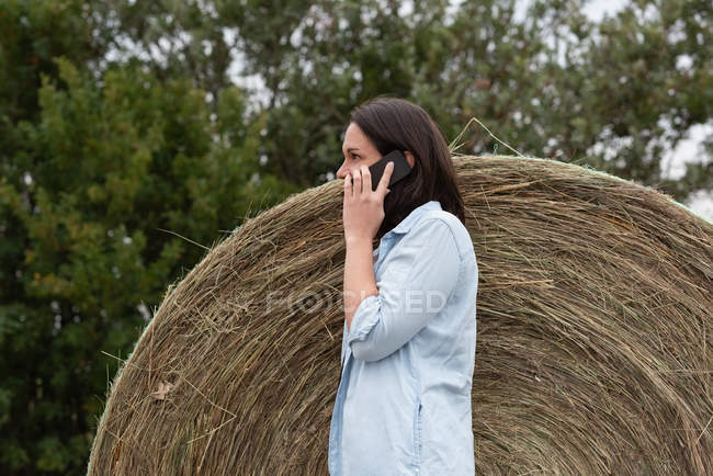 Vista lateral de la mujer hablando en el teléfono móvil mientras se apoya en la paca de heno - foto de stock