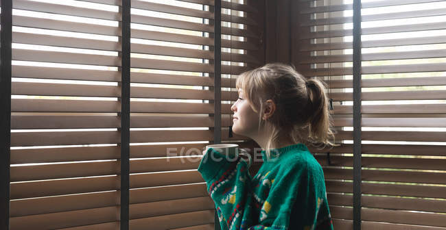 Vue latérale de la femme regardant par la fenêtre tout en prenant du café à la maison — Photo de stock