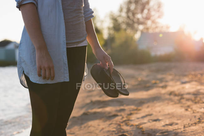 Sezione centrale della donna che tiene le pantofole sulla spiaggia — Foto stock
