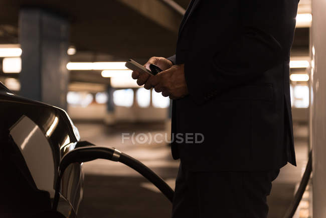 Uomo d'affari che utilizza il telefono cellulare durante la ricarica di auto elettriche alla stazione di ricarica — Foto stock