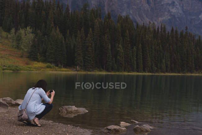 Rückansicht einer Frau, die Fotos mit Kamera am Seeufer anklickt — Stockfoto