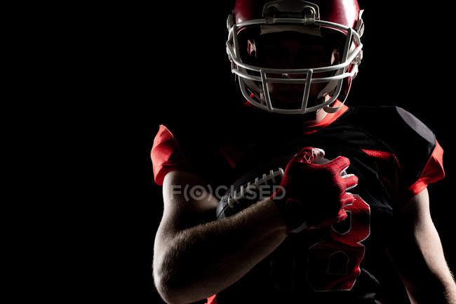 Giocatore di football americano in casco in possesso di palla da rugby contro sfondo nero — Foto stock