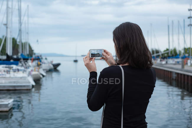 Вид ззаду жінки, що клацає фотографіями з мобільним телефоном біля гавані — стокове фото