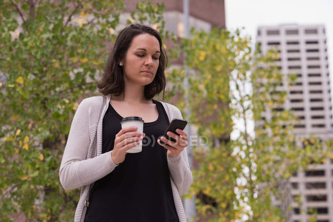 Mujer tomando café mientras usa el teléfono móvil en la ciudad - foto de stock