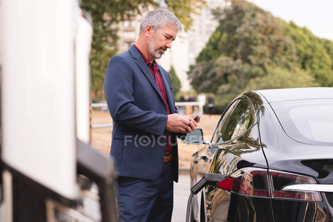Homme d'affaires utilisant un téléphone portable tout en rechargeant une voiture électrique par une journée ensoleillée — Photo de stock