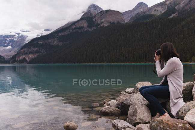 Vista laterale della donna cliccando foto con fotocamera vicino al lago — Foto stock