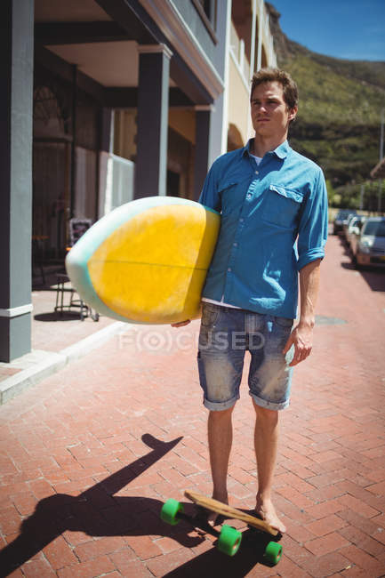 Man carrying surfboard while riding skateboard though footpath — Stock Photo
