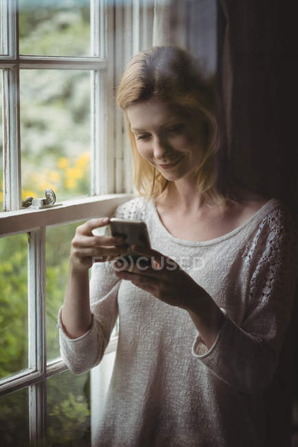 Mulher bonita de pé na janela e usando telefone celular em casa — Fotografia de Stock