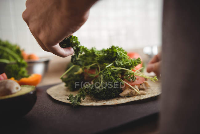 Gros plan des mains masculines plaçant des herbes sur le burrito sur le plan de travail de la cuisine — Photo de stock