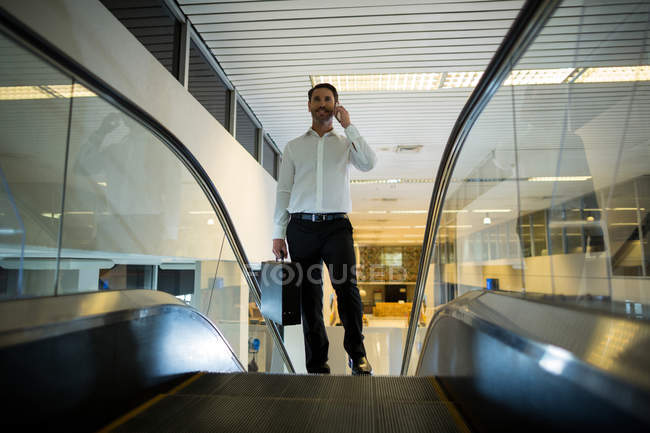 Hombre sonriente con maleta hablando por teléfono móvil en la escalera mecánica en el aeropuerto - foto de stock