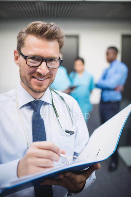 Retrato del médico sonriente escribiendo un informe médico en el pasillo del hospital - foto de stock