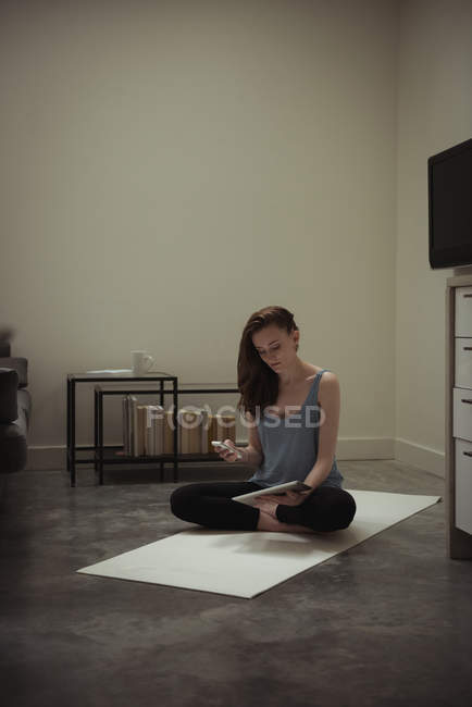 Здорова жінка використовує мобільний телефон і цифровий планшет на тренувальному килимку вдома — стокове фото