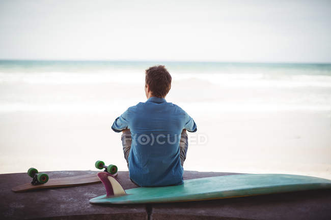 Вид сзади человека со скейтбордом и доской для серфинга, сидящего на пляже — стоковое фото