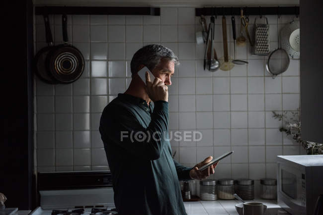 Чоловік розмовляє по телефону і використовує планшет на кухні — стокове фото