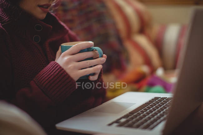A meio da seção da mulher que toma café ao usar o laptop no sofá — Fotografia de Stock