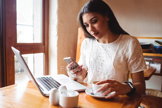 Schöne Frau benutzt Handy, während sie eine Tasse Kaffee im Café trinkt — Stockfoto