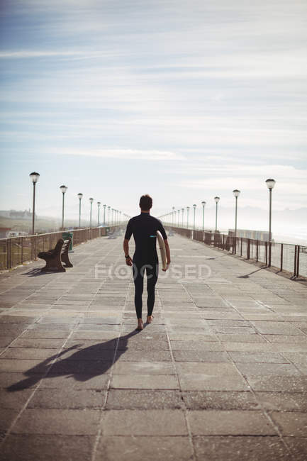 Vista posteriore del surfista che cammina con la tavola da surf sul molo in spiaggia — Foto stock
