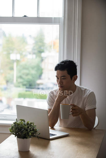 Uomo che guarda il computer portatile mentre prende una tazza di caffè a casa — Foto stock