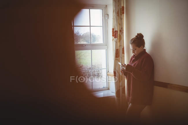 Mujer usando teléfono móvil cerca de la ventana en casa - foto de stock
