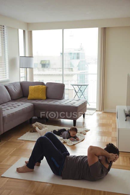 Padre haciendo ejercicio mientras el bebé juega en segundo plano en casa - foto de stock