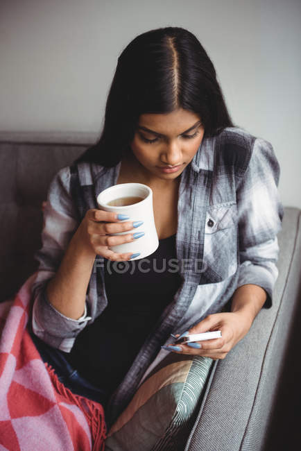 Mulher usando telefone celular enquanto segurando xícara de café na sala de estar em casa — Fotografia de Stock