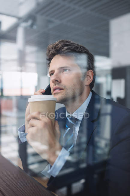 Бизнесмен, пользующийся мобильным телефоном и держащий в офисе одноразовую чашку кофе — стоковое фото
