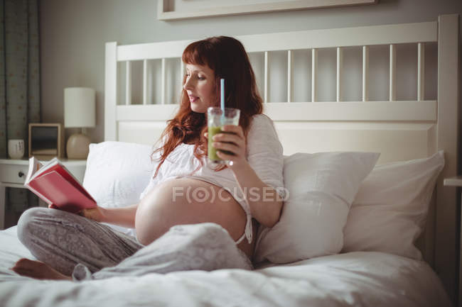 Вагітна жінка має сік під час читання книги на ліжку в спальні — стокове фото