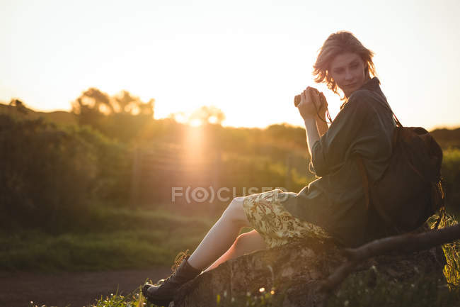 Mujer pensativa sentada en la roca y sosteniendo la cámara digital en un día soleado - foto de stock