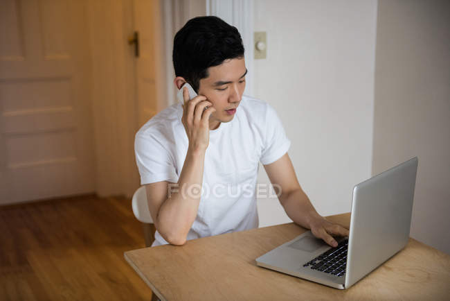 Hombre usando el ordenador portátil mientras habla en el teléfono móvil en casa - foto de stock