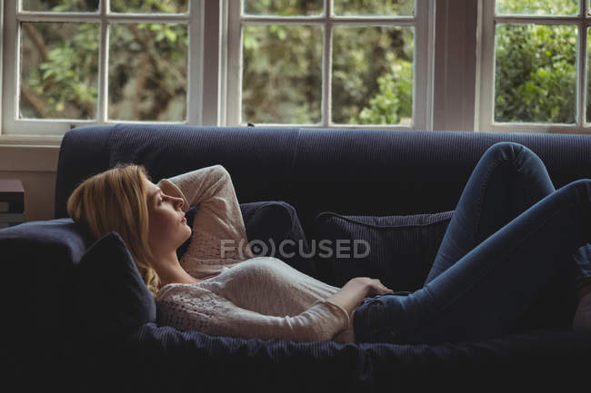 Mujer reflexiva acostada en el sofá en la sala de estar en casa - foto de stock
