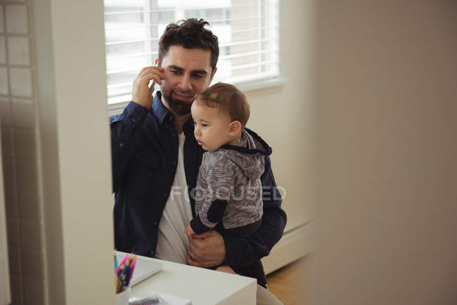 Батько розмовляє по мобільному телефону, тримаючи дитину вдома — стокове фото