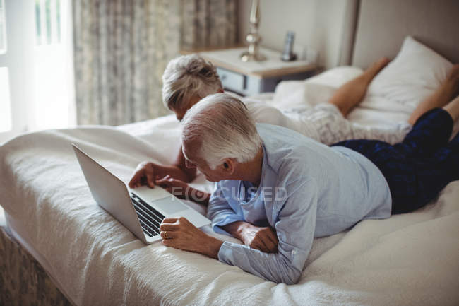 Старшая пара лежит на кровати и с помощью ноутбука в спальне — стоковое фото