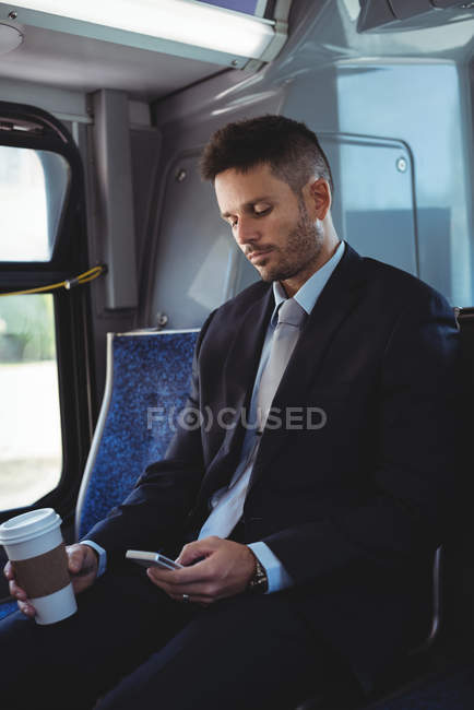Empresario sosteniendo una taza de café desechable y utilizando el teléfono móvil en el autobús - foto de stock