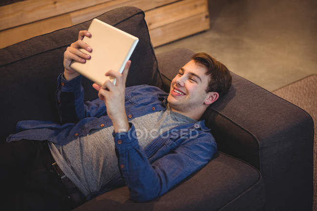 Hombre sonriente acostado en el sofá usando tableta digital en la sala de estar en casa - foto de stock
