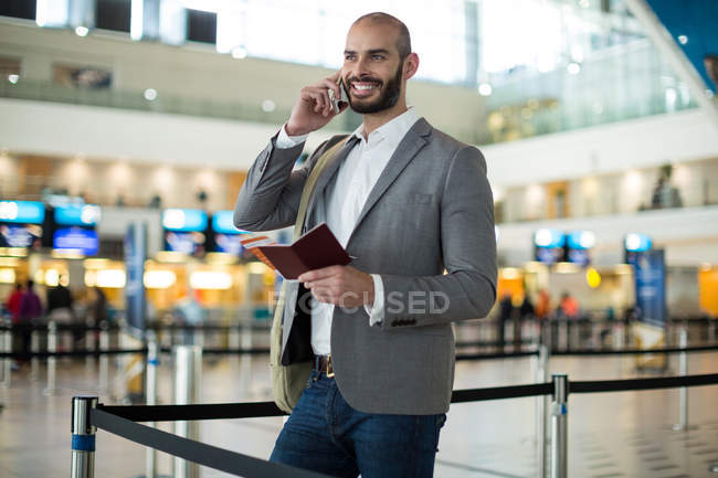 Улыбающийся бизнесмен, держащий посадочный талон и разговаривающий по мобильному телефону в терминале аэропорта — стоковое фото
