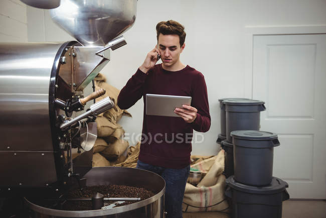 Мужчина разговаривает по мобильному телефону и с помощью цифрового планшета, стоя рядом с кофеваркой — стоковое фото