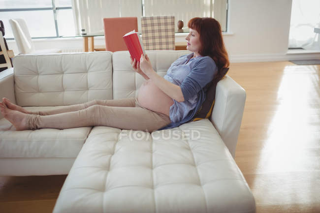 Pregnant woman reading book on sofa in living room at home — Stock Photo