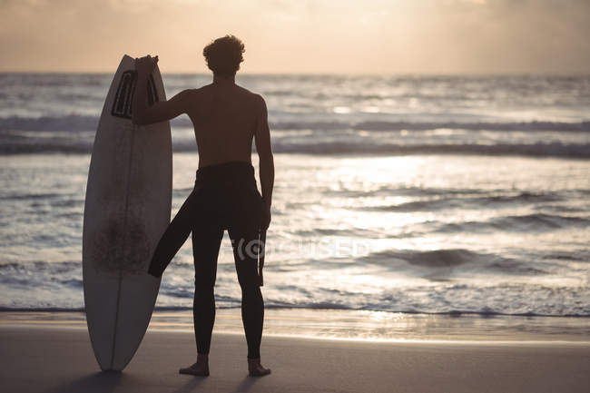 Vue arrière d'un homme portant une planche de surf debout sur la plage au crépuscule — Photo de stock