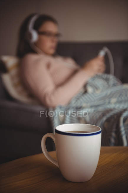 Xícara de café na mesa, enquanto a mulher ouvindo música no fundo em casa — Fotografia de Stock