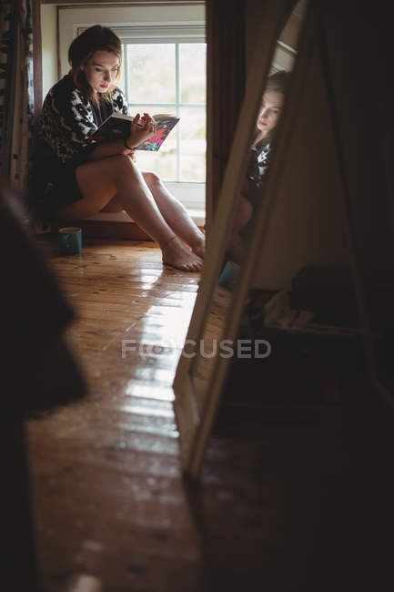 Frau sitzt auf Fensterbank und liest zu Hause ein Buch — Stockfoto