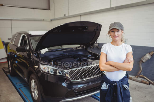 Female mechanic standing with arms crossed in front of car at repair garage — Stock Photo