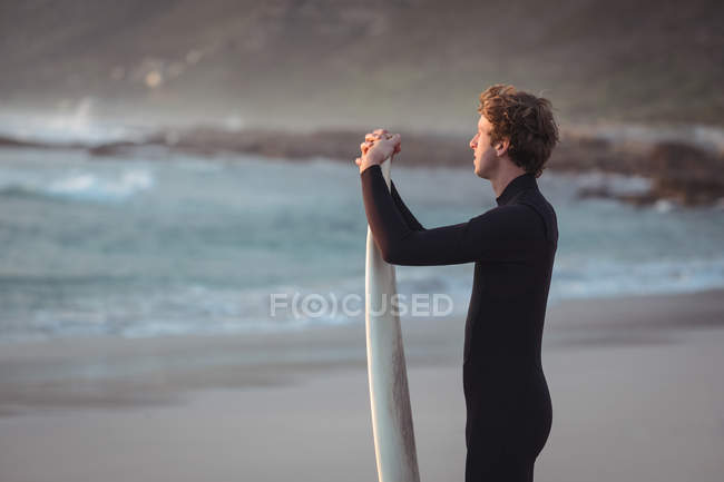 Вид збоку чоловіка в гідрокостюмі стоїть на пляжі з дошкою для серфінгу — стокове фото