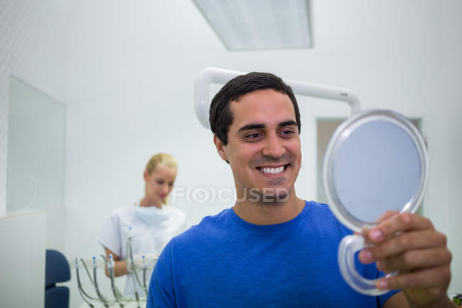 Patient checking teeth in mirror at dental clinic with female doctor in background — Stock Photo