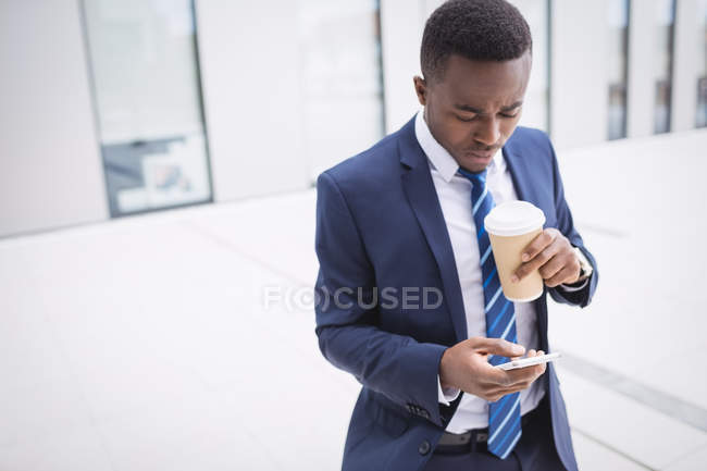 Empresário segurando copo de café descartável e usando telefone celular fora do prédio de escritórios — Fotografia de Stock